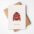 Kerstkaart met illustratie van een rode en groene kersttrui met rendiermotief en de tekst "warme wensen," liggend op een bruine envelop.