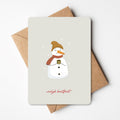 Kerstkaart met illustratie van een sneeuwpop met muts en sjaal, die een warme drank vasthoudt, omringd door sneeuwvlokken en de tekst "vrolijk kerstfeest," liggend op een bruine envelop.