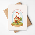 Een warm kaartje met een illustratie van een koppel dat samen hun nieuwe huis bewondert, onder een glazen stolp vol hartjes. De tekst ‘Gefeliciteerd met jullie nieuwe thuis’ maakt dit ontwerp perfect om iemand te feliciteren met een verhuis of nieuw hoofdstuk.
